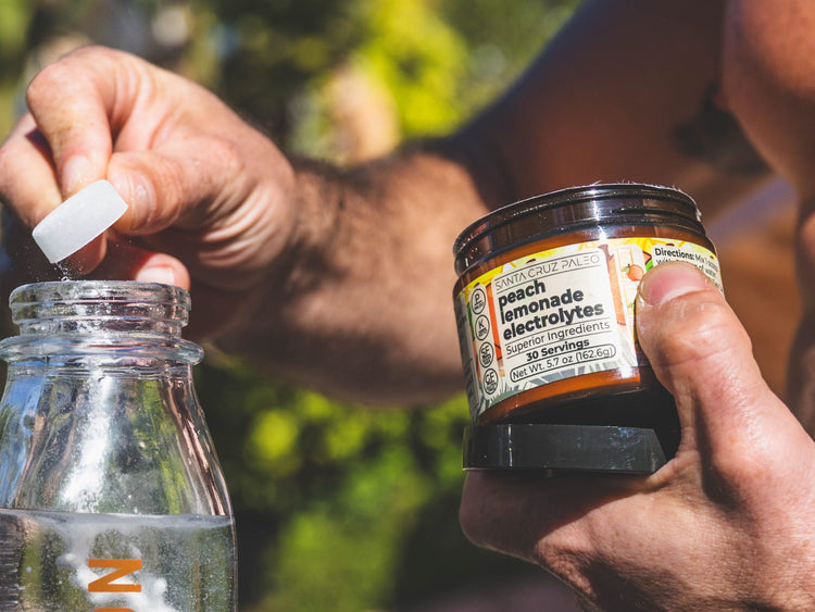 Hand opening a Santa Cruz Paleo Peach Lemonade electrolytes tub next to a glass water bottle