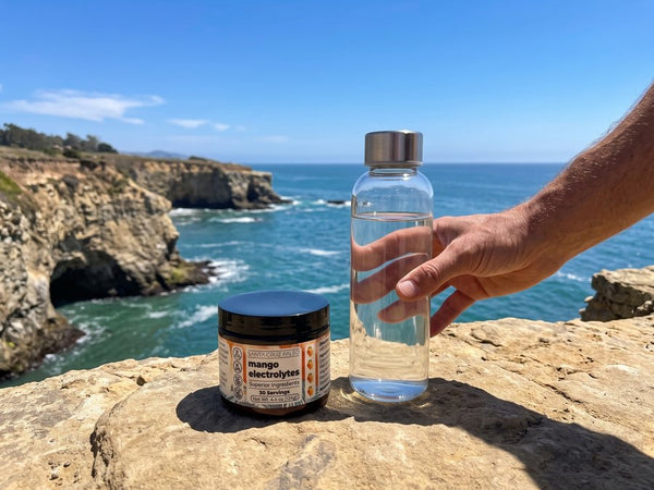 A hand reaching for the glass water bottle beside the Mango tub on a sea cliff